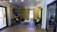 This single-floor space makes a strong design statement through its thoughtfully curated lounge area. The room is anchored by olive green walls that create a warm, energetic backdrop, complemented by rich hardwood flooring that transitions seamlessly into a bold navy and white geometric-patterned area rug. A set of modern charcoal swivel lounge chairs, accented with lime green throw pillows, are arranged around a sleek white tufted ottoman, creating an inviting and functional seating arrangement ideal for conversation or relaxation. A large abstract canvas artwork adorns one wall, adding an artistic focal point to the space. Natural light filters in through a centrally positioned window dressed with patterned curtains, brightening the room and providing a connection to the outdoors. Overhead, a track lighting system runs the length of the ceiling, offering adjustable, focused illumination throughout. Polished arc floor lamps further enhance the ambient lighting. The angled ceiling detail on one side of the room adds architectural character, while the overall layout feels open and well-proportioned. The space exudes a contemporary, boutique-like atmosphere with a confident blend of color, texture, and modern furnishings.