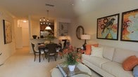 This single-floor residence showcases an open-concept layout that connects the living, dining, and kitchen areas into one cohesive, flowing space. The living area is anchored by a comfortable neutral sofa accented with warm orange and green throw pillows, complemented by a round glass coffee table adorned with vibrant floral arrangements. A framed decorative artwork and an oval mirror add visual interest to the walls, while a stylish table lamp contributes to the room's warm ambiance. The dining area transitions naturally from the living space and features a dark wood dining table surrounded by upholstered chairs beneath a modern multi-armed pendant light fixture. Just beyond, the kitchen is partially visible and includes dark wood cabinetry, stainless steel appliances, and a breakfast bar that reinforces the open flow between spaces. A built-in display niche provides additional architectural character. Throughout, the elevated ceilings enhance the sense of spaciousness, while the warm, neutral color palette is thoughtfully layered with earthy tones and organic accents, creating an inviting and well-appointed interior.