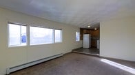 This single-floor space offers a straightforward, functional layout that connects a carpeted living area to a distinct kitchen zone. The living area is lined with large windows along one wall, allowing natural light to fill the room and providing views of the surrounding wooded landscape. A baseboard heating system runs beneath the windows, ensuring warmth throughout the space. The ceiling features a textured tile pattern that adds a subtle visual detail overhead.

A few steps down from the living area leads to the kitchen, which is set on a tiled floor and separated by a slight level change that creates a natural division between the two zones. The kitchen is equipped with warm-toned wood cabinetry, a white refrigerator, and additional appliances. The overhead lighting in the kitchen provides focused illumination for the workspace.

The overall ambiance of the space is practical and utilitarian, with neutral wall tones and a muted color palette throughout. The combination of carpeted and hard-surface flooring across the two areas helps define their respective functions. Large windows ensure the living space remains bright, while the connected kitchen layout supports convenient day-to-day use. This space is well-suited for those seeking a simple, no-frills environment with all essential areas within easy reach.