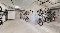 Located at 7 Erskine Ln in Victoria, BC, this dedicated bicycle storage room offers a practical and well-organized space for residents who rely on cycling as their primary mode of transportation. The room is constructed with exposed concrete walls and a textured concrete ceiling, giving it a utilitarian, industrial character that prioritizes function and durability.

The layout spans a generous, L-shaped floor plan that accommodates a substantial number of bicycles simultaneously. Wall-mounted bike hooks line multiple walls, allowing bicycles to be stored vertically — an efficient solution that maximizes the available floor space. The hooks are fitted with orange security cables, providing built-in locking points to help residents secure their bikes. Both standard bicycles and e-bikes can be observed making use of the storage system.

Overhead, exposed pipes and ventilation ductwork run along the ceiling, consistent with the room's no-frills, infrastructure-forward design. Lighting is provided by ceiling-mounted fixtures that ensure the space is well-lit and easy to navigate. A solid access door with a small window provides secure entry and exit.

The polished concrete floor is easy to maintain and well-suited to the demands of a high-traffic storage area. Overall, this bike storage room is a thoughtfully designed amenity that supports an active, urban lifestyle in Victoria's cycling-friendly community.