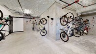 Located at 7 Erskine Ln in Victoria, BC, this dedicated bicycle storage room offers a practical and well-organized space for residents who rely on cycling as their primary mode of transportation. The room is constructed with exposed concrete walls and a textured concrete ceiling, giving it a utilitarian, industrial character that prioritizes function and durability.

The layout spans a generous, L-shaped floor plan that accommodates a substantial number of bicycles simultaneously. Wall-mounted bike hooks line multiple walls, allowing bicycles to be stored vertically — an efficient solution that maximizes the available floor space. The hooks are fitted with orange security cables, providing built-in locking points to help residents secure their bikes. Both standard bicycles and e-bikes can be observed making use of the storage system.

Overhead, exposed pipes and ventilation ductwork run along the ceiling, consistent with the room's no-frills, infrastructure-forward design. Lighting is provided by ceiling-mounted fixtures that ensure the space is well-lit and easy to navigate. A solid access door with a small window provides secure entry and exit.

The polished concrete floor is easy to maintain and well-suited to the demands of a high-traffic storage area. Overall, this bike storage room is a thoughtfully designed amenity that supports an active, urban lifestyle in Victoria's cycling-friendly community.