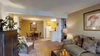 This single-floor residence offers an open-concept layout that seamlessly connects the living, dining, and kitchen areas into one cohesive living space. The living area is carpeted and comfortably accommodates a sofa and wicker accent chair, with warm lighting provided by a decorative table lamp. A glass-top dining table with wrought iron chairs sits nearby, lit by a pendant light overhead and accented by framed botanical wall art, adding a classic touch to the space.

The kitchen is partially open to the main living area, separated by a white breakfast bar that provides additional counter space and casual seating options. Warm wood-toned cabinetry lines the kitchen walls, complemented by white appliances including a refrigerator. Recessed fluorescent lighting illuminates the kitchen work area effectively. A structural column near the kitchen entry adds architectural definition between the spaces.

Throughout the residence, walls are finished in warm golden-beige tones that create a cozy, inviting ambiance. The consistent carpeting across the main living and dining areas contributes to a unified, comfortable feel throughout this well-organized single-level home.