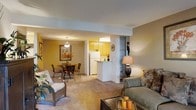 This single-floor residence offers an open-concept layout that seamlessly connects the living, dining, and kitchen areas into one cohesive living space. The living area is carpeted and comfortably accommodates a sofa and wicker accent chair, with warm lighting provided by a decorative table lamp. A glass-top dining table with wrought iron chairs sits nearby, lit by a pendant light overhead and accented by framed botanical wall art, adding a classic touch to the space.

The kitchen is partially open to the main living area, separated by a white breakfast bar that provides additional counter space and casual seating options. Warm wood-toned cabinetry lines the kitchen walls, complemented by white appliances including a refrigerator. Recessed fluorescent lighting illuminates the kitchen work area effectively. A structural column near the kitchen entry adds architectural definition between the spaces.

Throughout the residence, walls are finished in warm golden-beige tones that create a cozy, inviting ambiance. The consistent carpeting across the main living and dining areas contributes to a unified, comfortable feel throughout this well-organized single-level home.