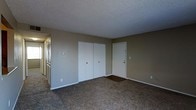 This single-floor residential space offers a straightforward and functional layout designed to maximize usability. The main room is finished with wall-to-wall carpeting in a neutral tone, providing a warm and comfortable underfoot feel throughout. The walls are painted in a muted beige, lending a calm and understated ambiance to the interior.

A prominent feature of the room is the set of double bi-fold closet doors, offering generous built-in storage that keeps the living area organized and uncluttered. A solid entry door with a standard door viewer is positioned nearby, providing direct access to the space. On the opposite side, a row of windows allows natural light to filter in, brightening the interior and offering a connection to the outdoors.

The textured popcorn ceiling adds a classic residential character to the space. Clean white trim and baseboards run along the perimeter of the room, providing a crisp contrast against the neutral walls and flooring. Electrical outlets and light switch plates are conveniently positioned throughout the room.

Overall, this space presents a clean, move-in ready environment with a no-frills, practical design that suits a variety of uses. The neutral color palette and simple finishes create a versatile backdrop, easily adaptable to a range of furnishing styles and personal tastes.
