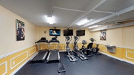 This single-floor fitness room offers a focused cardio workout environment equipped with essential exercise machines. The room houses two treadmills, two elliptical trainers, and a stationary bike, all arranged along the perimeter to maximize usable floor space. Three wall-mounted flat-screen televisions are positioned at eye level, allowing users to stay entertained during workouts. The flooring consists of durable dark rubber matting, providing both traction and impact absorption suitable for exercise use. The walls are painted in a warm yellow tone with white wainscoting-style paneling, lending the space a bright and welcoming feel. Overhead fluorescent strip lighting ensures the room is well-illuminated throughout. Exposed white piping runs along the ceiling, adding an industrial touch to the otherwise clean aesthetic. A motivational fitness poster adorns one wall, reinforcing the room's athletic purpose. The layout is efficiently organized, making the most of the available space while keeping equipment accessible and the environment functional for daily cardio training.