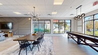 Located at 21 East St in North Attleboro, MA, this single-floor amenity space is thoughtfully designed to offer a modern, welcoming environment for gathering, socializing, and recreation. The open-plan layout flows seamlessly between distinct functional zones, all unified by light hardwood-style flooring, a drop tile ceiling, and abundant natural light streaming through large, black-framed windows that offer views of the surrounding greenery.

The lounge area serves as the heart of the space, anchored by a rich cognac leather sofa, stylish accent chairs, and a striking geometric accent wall finished in soft gray with angular panel detailing. A large flat-screen television is mounted to this feature wall, making it an ideal spot for relaxed viewing. Pendant lighting and an eye-catching sputnik chandelier add architectural character throughout.

Adjacent to the lounge, a generously sized dining table with seating for twelve sits beneath a bold starburst chandelier, creating an inviting setting for communal meals or events. A billiards table and shuffleboard round out the entertainment offerings in the game area nearby.

The fully equipped kitchen features sleek gray cabinetry, granite countertops, stainless steel appliances including a wall oven, side-by-side refrigerator, and dishwasher — ideal for catering or everyday use. The overall ambiance is contemporary, clean, and versatile.