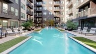 Located at 905 S Harwood St in Dallas, TX, this outdoor courtyard amenity space is thoughtfully designed to serve as a central gathering hub within a modern multi-story residential community. The space is organized around a large, rectangular pool with striking blue water, flanked by turf-lined decks furnished with chaise lounges and wicker seating arrangements, creating a resort-like atmosphere in the heart of the city.

A bold black steel pergola anchors one section of the courtyard, providing partial shade over an intimate lounge area outfitted with cushioned wicker sofas and coffee tables. String lights are strung overhead in adjacent seating zones, adding a warm, inviting ambiance for evening gatherings. Brick-patterned hardscape pathways connect the various areas, complemented by well-maintained artificial turf sections and mature landscaping that adds natural texture and greenery throughout.

The surrounding multi-story building facades, finished in charcoal siding with large windows and private balconies, frame the courtyard on all sides, offering a sense of enclosure and community. Raised planter beds and ornamental trees soften the architecture, while thoughtfully placed outdoor lighting ensures the space remains functional and attractive day and night.