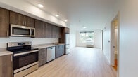 Located at 510 NE Dekum St in Portland's vibrant Northeast neighborhood, this single-floor unit offers a clean, contemporary design throughout. Light wood-tone flooring flows seamlessly across the open-concept layout, connecting the kitchen and living areas in a cohesive, airy arrangement.

The kitchen is appointed with rich dark wood cabinetry extending floor to ceiling, providing ample storage. A full suite of stainless steel appliances includes an electric range, over-the-range microwave, dishwasher, and refrigerator, all complemented by light-toned countertops and a stainless steel sink. A dedicated nook houses a stacked in-unit washer and dryer for added convenience.

The spacious living area is bathed in natural light through large windows that frame views of the surrounding neighborhood's mature tree canopy. A sliding barn door adds a modern architectural accent, while a wall-mounted HVAC unit ensures year-round comfort. The bedroom is a bright, minimalist space with clean white walls and generous natural light.

The bathroom features a full soaking tub and shower combination with chrome fixtures and a curved curtain rod, finished in crisp white surroundings. Recessed lighting throughout the unit adds to the modern, polished ambiance, making this a well-appointed and functional Portland residence.