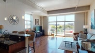 This stylish single-floor residence offers a thoughtfully designed open-concept layout that seamlessly connects the living, dining, and kitchen areas. Rich hardwood flooring flows throughout the main living space, complemented by soaring ceilings with industrial-style track lighting. Expansive floor-to-ceiling windows flood the interior with abundant natural light and frame sweeping outdoor city views, while a glass-railed balcony provides direct access to the outdoors — ideal for enjoying the surrounding urban landscape.

The kitchen is a functional centerpiece, featuring warm wood cabinetry, dark granite countertops, a mosaic tile backsplash, and a full suite of stainless steel appliances. A breakfast bar with upholstered seating creates a natural gathering point between the kitchen and living area.

The living room is generously proportioned, with a cozy seating arrangement and large-format artwork that adds character to the neutral walls. The bedroom is a calm retreat with plush carpeting, a ceiling fan, and a window with city views. The bathroom is finished with tile flooring, a dark granite vanity with wood cabinetry, a large mirror, and a walk-in shower, offering a clean and polished aesthetic throughout.