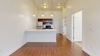 Located at 147 N Harold St in Salt Lake City, Utah, this single-floor residence offers a thoughtfully designed open-concept layout that seamlessly connects the kitchen and living areas. The kitchen is a standout feature, showcasing rich dark-stained cabinetry that extends toward dramatically high ceilings, stainless steel appliances including a refrigerator, electric range, and built-in microwave, as well as a dishwasher. A peninsula-style island with a double stainless steel sink provides additional prep space and is accented by a trio of elegant pendant lights hanging overhead, enhancing the sense of vertical space. The open living area benefits from abundant natural light streaming in through French doors fitted with blinds, which also offer direct access to the outdoors. Warm hardwood-style flooring runs throughout the main living spaces, adding a cohesive and inviting feel. The layout also includes a separate laundry area and additional rooms accessible via interior doors, offering functional flexibility. The bright, neutral white walls and generous ceiling heights throughout contribute to an airy, open ambiance that makes the space feel expansive and welcoming.