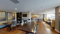 This single-floor communal space is thoughtfully designed to accommodate a variety of activities across several well-defined zones. The dining area anchors one end of the room, featuring large dark-wood tables paired with mixed seating and hardwood flooring that extends throughout much of the space. A central focal point is the tile-surround fireplace topped with a wall-mounted flat-screen TV and flanked by dark built-in shelving, creating a sophisticated media and lounge hub.

Adjacent to the fireplace area, a casual seating zone is furnished with a neutral sofa, a game table, and patterned armchairs, accented by a large multi-panel abstract artwork in warm amber and grey tones. A curved window alcove at the far end offers an abundance of natural light through floor-to-ceiling windows dressed with sheer vertical drapes, furnished with gold-upholstered wingback chairs, tufted ottomans, and a sleek side table—ideal for quiet reading or conversation.

The kitchenette features dark cabinetry, stone-look countertops, stainless steel appliances including a refrigerator and coffee station, and warm wood-look flooring. Recessed lighting throughout ensures the entire space remains bright and welcoming, while curated artwork and greenery contribute to a polished, contemporary ambiance.