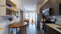 This well-appointed single-floor residence offers a thoughtfully designed open-concept layout that seamlessly connects the kitchen, dining, and living areas. The kitchen is a standout feature, showcasing rich dark-wood cabinetry, granite countertops, a white subway tile backsplash, stainless steel appliances including a refrigerator, range, microwave, and dishwasher, and a double-basin sink with a modern gooseneck faucet. A cozy dining nook with a round wood table sits adjacent to the kitchen, complemented by a decorative wine rack wall feature. The living and bedroom area is generously proportioned, bathed in natural light through large floor-to-ceiling windows with vertical blinds that open to an outdoor view. Elevated ceilings and a ceiling fan enhance the sense of airiness throughout. Wood-style plank flooring runs continuously across the space, grounding the contemporary aesthetic. The bathroom is finished with a granite-topped dark wood vanity, an oversized mirror, and direct access to a spacious walk-in closet with built-in rod storage. Neutral tones, clean lines, and modern finishes throughout create a cohesive and functional living environment.