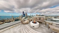 This stunning 870 square foot space in Seattle showcases contemporary design and breathtaking views. The interior boasts an open floor plan with floor-to-ceiling windows that flood the area with natural light and offer panoramic vistas of the city skyline and water. A striking feature is the wooden slatted ceiling, adding warmth and texture to the modern aesthetic.

The living area is anchored by a sleek, two-sided fireplace, creating a cozy atmosphere. Plush seating, including vibrant orange armchairs and a curved white sofa, provides comfortable lounging spaces. A minimalist kitchen with a central island integrates seamlessly into the layout.

Multiple outdoor terraces extend the living space, featuring glass railings for unobstructed views. These areas are furnished with modern outdoor seating, perfect for relaxation or entertainment. The property's elevation allows for sweeping views of the urban landscape and nearby water, making it an ideal spot for both day and night enjoyment.

The thoughtful design combines functionality with luxury, evident in details like the built-in storage and carefully chosen furniture pieces. This space epitomizes urban sophistication, offering a tranquil retreat high above the bustling city below.