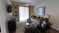 This single-floor residence centers around a thoughtfully designed living room that balances comfort and contemporary style. The space is finished in warm neutral tones throughout, with beige walls providing a versatile backdrop for the carefully selected furnishings and décor. A ceiling fan with integrated lighting serves as a functional focal point overhead, promoting airflow and ambiance.

The living room features a plush, light-toned sofa adorned with decorative pillows in earthy oranges and deep blacks, complemented by a coordinating armchair with patterned cushions. Two sleek, glass-topped coffee tables are arranged in a staggered layout on a stone-tile floor, adding a modern touch to the space. A leather ottoman anchors the seating arrangement at the front of the room.

Large framed artwork — including a prominent cityscape print — decorates the walls alongside a curated gallery arrangement of three smaller framed pieces. A floor-to-ceiling roller shade covers a wide sliding glass door, allowing filtered natural light to brighten the interior while providing privacy. Drapes frame the window, adding texture and warmth. The overall ambiance is polished, inviting, and move-in ready.