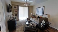 This single-floor residence centers around a thoughtfully designed living room that balances comfort and contemporary style. The space is finished in warm neutral tones throughout, with beige walls providing a versatile backdrop for the carefully selected furnishings and décor. A ceiling fan with integrated lighting serves as a functional focal point overhead, promoting airflow and ambiance.

The living room features a plush, light-toned sofa adorned with decorative pillows in earthy oranges and deep blacks, complemented by a coordinating armchair with patterned cushions. Two sleek, glass-topped coffee tables are arranged in a staggered layout on a stone-tile floor, adding a modern touch to the space. A leather ottoman anchors the seating arrangement at the front of the room.

Large framed artwork — including a prominent cityscape print — decorates the walls alongside a curated gallery arrangement of three smaller framed pieces. A floor-to-ceiling roller shade covers a wide sliding glass door, allowing filtered natural light to brighten the interior while providing privacy. Drapes frame the window, adding texture and warmth. The overall ambiance is polished, inviting, and move-in ready.