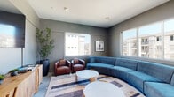 This single-floor communal lounge space in Modesto, California, is designed with a contemporary aesthetic that balances comfort and function. The room is anchored by a large, curved blue sectional sofa positioned at the center, complemented by a pair of rich brown leather club chairs arranged around a set of round marble-top coffee tables. The seating arrangement rests atop a geometric, multi-toned area rug that adds visual interest to the floor.

The walls are finished in a cool, medium-toned gray, contributing to the space's modern, relaxed atmosphere. Recessed lighting in the white ceiling provides consistent illumination throughout. Multiple large windows along two walls flood the room with natural light and offer views of the surrounding community buildings. Framed black-and-white artwork accents the walls, adding a curated, polished touch.

A natural wood credenza along one wall supports a wall-mounted flat-screen television, with decorative books and small plants styled on its surface. Tall potted greenery in a cylindrical planter adds a natural element to the otherwise clean-lined interior. Glass-paned doors adjacent to the lounge provide direct access to a well-equipped fitness center, seamlessly connecting the two amenity spaces.