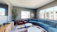 This single-floor communal lounge space in Modesto, California, is designed with a contemporary aesthetic that balances comfort and function. The room is anchored by a large, curved blue sectional sofa positioned at the center, complemented by a pair of rich brown leather club chairs arranged around a set of round marble-top coffee tables. The seating arrangement rests atop a geometric, multi-toned area rug that adds visual interest to the floor.

The walls are finished in a cool, medium-toned gray, contributing to the space's modern, relaxed atmosphere. Recessed lighting in the white ceiling provides consistent illumination throughout. Multiple large windows along two walls flood the room with natural light and offer views of the surrounding community buildings. Framed black-and-white artwork accents the walls, adding a curated, polished touch.

A natural wood credenza along one wall supports a wall-mounted flat-screen television, with decorative books and small plants styled on its surface. Tall potted greenery in a cylindrical planter adds a natural element to the otherwise clean-lined interior. Glass-paned doors adjacent to the lounge provide direct access to a well-equipped fitness center, seamlessly connecting the two amenity spaces.