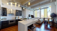Located within the Goldtex building on N 12th Street in Philadelphia, this single-floor loft-style residence showcases an industrial-modern design highlighted by soaring ceilings, exposed silver ductwork, and rich hardwood flooring throughout. The open-plan living area benefits from expansive floor-to-ceiling windows that flood the space with natural light while framing sweeping views of the Philadelphia skyline.

The kitchen is fitted with dark, wood-grain cabinetry, white quartz countertops, a stainless steel range hood, and built-in appliances, anchored by a generous island that doubles as a workspace and casual dining counter. The adjacent living and dining areas are generous and airy, illuminated by pendant lighting and warmed by the rich tones of the hardwood floors.

The bedroom features tall windows with urban views, mirrored sliding closet doors, and frosted glass doors adding privacy. Two bathrooms complete the unit—one with a bold teal accent wall, white vanity, and modern fixtures, and another featuring a soaking tub with a rainfall shower panel, floating white vanity, and wood-look tile flooring. Overall, the space offers a cohesive blend of industrial character and contemporary refinement.