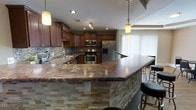 This single-floor residence showcases a thoughtfully designed open-concept layout centered around an impressive kitchen. The kitchen is a standout feature, boasting rich, dark-toned granite countertops with warm amber and burgundy tones that extend across a generously sized breakfast bar with stacked stone detailing — an elegant architectural element that adds texture and visual depth to the space. The bar area is furnished with sleek bar stools, making it ideal for casual dining or entertaining. Rich walnut-toned cabinetry lines the kitchen walls, providing ample storage, while a decorative tile backsplash adds a refined touch. The kitchen is fully equipped with a suite of stainless steel appliances, including a double wall oven, a gas range, and a French door refrigerator with freezer drawer. Recessed ceiling lighting and stylish pendant lights illuminate the workspace, creating a warm and inviting ambiance. Large vertical blinds along the adjacent window wall allow for flexible natural light control, brightening the open space. The overall design balances functionality with upscale finishes, resulting in a cohesive and polished interior aesthetic throughout this single-level home.