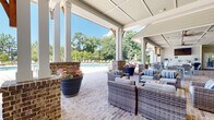 Located at 100 Pinckney Marsh Ln in Port Royal, South Carolina, this single-story amenity pavilion is designed to serve as a premier outdoor gathering and recreation destination. The structure's architecture blends Lowcountry charm with modern functionality, showcasing a generously proportioned covered patio defined by white-painted tongue-and-groove ceilings, exposed wood bracket accents, and sturdy brick column bases that lend a refined yet relaxed aesthetic.

The expansive brick-paved terrace flows seamlessly beneath the covered roof, accommodating multiple distinct activity zones. Comfortable wicker lounge seating with neutral cushions and striped accent pillows creates inviting conversation areas, while dining tables with upright chairs offer a more structured setting for meals and gatherings. A dedicated recreation area features a ping-pong table, adding a casual, social element to the space.

An outdoor kitchen and bar station with white cabinetry and an overhead mounted television serves the dining zone, making the pavilion well-suited for entertaining. Multiple ceiling fans are mounted throughout, ensuring comfortable airflow during warm South Carolina days.

The pavilion opens directly onto a large pool deck with lounge chairs and umbrellas, surrounded by mature trees that provide a natural, lush backdrop. The open-air design floods the space with abundant natural light and unobstructed views of the pool and surrounding landscape, creating a bright and welcoming atmosphere throughout.