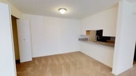This single-floor unit offers a straightforward and functional layout designed for comfortable everyday living. The main living area features wall-to-wall carpeting in a neutral beige tone, creating a warm and understated atmosphere that pairs well with the clean, white-painted walls throughout.

A defining architectural detail is the pass-through window that connects the living area to the kitchen, complete with a granite-style countertop and a sink visible from the main room. This open connection between spaces promotes a sense of flow and makes the layout feel more open and communicative. The kitchen area features dark cabinetry visible through the pass-through opening, providing a subtle contrast to the lighter tones of the surrounding space.

Ceiling-mounted flush lighting provides consistent illumination across the living area, keeping the space bright and evenly lit. The walls are smooth and freshly finished, offering a blank canvas ready for personalization. The overall ambiance is clean, minimal, and move-in ready, with a neutral color palette that suits a variety of interior styles. This unit is well-suited for those seeking a low-maintenance, efficiently designed space with a simple, uncluttered aesthetic.