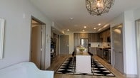 This single-floor residence showcases a thoughtfully designed open-concept layout that seamlessly connects the kitchen and dining areas into one cohesive living space. The kitchen is outfitted with dark wood cabinetry, stainless steel appliances including a refrigerator and gas range, and a clean white kitchen island that doubles as a breakfast bar. Recessed lighting illuminates the space efficiently, complemented by a striking geometric pendant light fixture above the dining area, which adds a bold decorative focal point. The dining space is anchored by a bold black-and-white diamond-patterned area rug, with a rectangular dining table and dark chairs arranged beneath the pendant light. Light wood-toned flooring runs throughout, adding warmth and continuity across the open floor plan. A hallway visible from the main living area leads to additional rooms and features multiple doors, suggesting a practical and organized layout. The overall design aesthetic blends contemporary and transitional elements, with contrasting dark cabinetry, light walls, and eclectic décor accents creating a visually dynamic yet welcoming atmosphere.