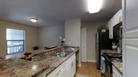 This single-floor residence offers a thoughtfully designed layout that balances comfort and functionality. The kitchen serves as a central highlight, showcasing rich, granite-style countertops that provide ample prep space alongside classic white cabinetry with brushed nickel hardware. A double-basin stainless steel sink is seamlessly integrated into the countertop, complemented by a full suite of stainless steel appliances, including a refrigerator and range. Warm hardwood flooring runs throughout, adding a welcoming and cohesive feel to the space. A textured ceiling adds subtle character overhead, while flush-mount lighting ensures the kitchen remains bright and well-lit. A closeted storage area is conveniently tucked nearby, offering practical organization solutions. The neutral, soft grey wall tones create a calm and versatile backdrop that allows for a variety of interior styles. The open sight lines between the kitchen and adjacent living areas enhance the sense of space, making the layout feel connected and easy to navigate. Overall, the residence presents a clean, move-in-ready aesthetic with durable finishes well-suited for everyday living.