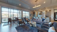 This single-floor clubhouse is designed with a modern aesthetic, offering a welcoming and well-appointed communal environment. The main lounge area is expansive and open, featuring wide-plank hardwood flooring, high ceilings, and an abundance of natural light streaming through large floor-to-ceiling windows and glass panel doors. These doors open to views of a covered outdoor terrace and pool area beyond, seamlessly connecting the interior to the exterior amenity spaces.

The lounge is furnished with plush sofas, upholstered armchairs, and area rugs, creating distinct seating zones suitable for relaxing or socializing. A sleek, built-in fireplace with a textured tile surround anchors one wall, accompanied by a wall-mounted television and curated artwork, adding warmth and visual interest to the space.

The clubhouse also includes a fully equipped kitchen, highlighted by a stainless steel range with a polished metal hood, a matching refrigerator, a reflective penny-tile backsplash, and a clean white island with bar seating. Globe pendant lights and a coffee station add functionality and character to this area.

A separate dining zone with round tables and chairs is positioned near the large windows, making it an ideal spot for casual meals or gatherings. Throughout, the design balances contemporary finishes with comfortable furnishings, creating an inviting atmosphere for residents to gather and connect.