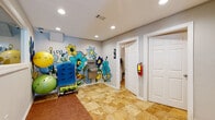 Located at 150 Chateau Terrace in Athens, Georgia, this yoga and wellness studio offers a thoughtfully designed single-floor space that balances functionality with an energizing, artistic atmosphere. The studio's most striking feature is a bold, hand-painted mural adorning the main lobby area — a vivid composition of oversized flowers in shades of blue, yellow, and green, accompanied by the words "Love" and "Meditate," instantly setting a calming yet uplifting tone for visitors.

The flooring throughout combines warm-toned stone tile with wood-panel exercise matting, creating distinct zones for movement and transition. Recessed lighting illuminates the space evenly, contributing to a clean and welcoming ambiance. A built-in water fountain station with bottle-filling capability is conveniently positioned in the lobby area alongside a rack of exercise balls and instructional fitness equipment.

An interior pass-through window connects the lobby to what appears to be a reception or administrative area, providing seamless communication between staff and guests. A hallway leads to accessible restrooms for both men and women, and a glass-paneled entry door allows natural light to filter into the corridor. The studio's layout is efficient and well-organized, designed to accommodate practitioners of all levels in a space that feels both professional and warmly inviting.