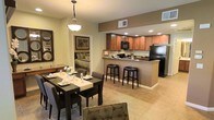 This single-floor space showcases an open-concept layout that seamlessly connects the kitchen, dining, and living areas. The kitchen is equipped with warm-toned wooden cabinetry, dark granite countertops, black appliances including a refrigerator and microwave, and recessed lighting. A raised breakfast bar with two cushioned bar stools provides a casual seating option and serves as a natural divider between the kitchen and dining area. The dining space features a dark rectangular table comfortably set for several guests, complemented by matching upholstered chairs and a classic pendant light overhead. Light ceramic tile flooring runs throughout, contributing to the bright, cohesive feel of the space. A glimpse through a doorway reveals a bedroom with a tufted headboard and coordinating furnishings, suggesting a well-appointed sleeping area. Neutral beige and taupe wall tones paired with warm cabinetry create an inviting and polished ambiance. The thoughtful layout maximizes functionality while maintaining a comfortable, well-furnished aesthetic suited for both everyday living and entertaining.