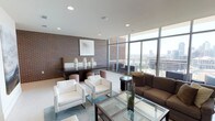 This stylish lounge space in Dallas offers an impressive blend of urban sophistication and contemporary design. The single-floor layout is anchored by dramatic floor-to-ceiling glass windows that frame sweeping panoramic views of the Dallas skyline, flooding the interior with abundant natural light throughout the day.

Exposed brick walls serve as a defining architectural element, adding warmth and texture that contrasts beautifully with the clean white ceilings and smooth flooring. The space is divided into distinct seating areas, each thoughtfully arranged for comfort and conversation. A lounge area features crisp white armchairs and a glass-topped coffee table set atop a teal area rug, while a separate seating zone offers a plush brown sofa with coordinating accent pieces.

A media area is built into a rich crimson accent wall, housing a large flat-screen television above dark wood cabinetry and flanked by large-scale abstract artwork on the adjacent brick wall. A communal dining or gathering table with upholstered bench seating and woven ottoman stools provides additional functionality, adorned with sculptural white ceramic vases. The overall ambiance is polished and modern, making it an inviting and versatile space well-suited for both relaxation and socializing.