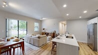 This single-floor residence at Natura Gardens offers a well-designed, open-concept layout with a seamless flow between the living, dining, and kitchen areas. Light-toned walls and light wood-look flooring run throughout, creating a bright and cohesive aesthetic.

The kitchen is a functional centerpiece, outfitted with sleek white cabinetry extending to the ceiling for ample storage, a full suite of stainless steel appliances including a side-by-side refrigerator, electric range, and built-in microwave, and a quartz countertop island with an integrated sink that also serves as a breakfast bar. The adjacent living area features large windows that invite natural light and offer views of the surrounding greenery. A separate dining space accommodates a full dining set beneath track lighting.

The bedrooms are comfortably sized with natural light from windows and include closet storage. One bedroom features a walk-in style closet with built-in shelving and hanging space. The bathroom is finished with a quartz vanity countertop, a large wall mirror, and a shower-tub combination with marble-style tile surround.

Overall, the space presents a clean, modern atmosphere with consistent finishes and a practical floor plan suited for comfortable everyday living.