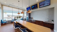 This contemporary amenity lounge is thoughtfully designed to offer both functionality and comfort. The open-plan layout is anchored by a generously sized kitchen bar with a warm wood-toned surface, complemented by a row of sleek bar stools in navy blue. Suspended above the island, a trio of copper pendant lights adds a striking design element and warm ambiance to the space.

The kitchen wall features rich dark wood cabinetry, a textured mosaic tile backsplash, and built-in appliances including a beverage center and dishwasher drawer. Multiple flat-screen televisions are mounted above for entertainment. Light wood-toned flooring flows throughout, tying the various zones together seamlessly.

Floor-to-ceiling windows and clerestory glazing flood the interior with abundant natural light while offering panoramic views of the surrounding cityscape. A cozy lounge area is defined by a bold deep-blue accent wall adorned with a curated collection of circular macramé wall art. Plush seating includes a neutral linen sofa and warm cognac leather chairs, creating an inviting retreat.

Glass-panel doors open directly to an outdoor terrace, visually expanding the space and blurring the boundary between indoors and out. A windowed nook with built-in banquette seating and small bistro tables provides an ideal spot for intimate gatherings. Recessed lighting throughout completes this well-appointed, design-forward communal space.