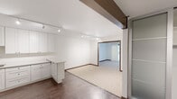 This single-floor space offers a clean, modern aesthetic with a thoughtful layout that balances open living areas with defined private spaces. The kitchen is a standout feature, outfitted with crisp white shaker-style cabinetry extending to the ceiling for ample storage, light-toned countertops, a stainless steel sink, and a full suite of stainless steel appliances including a refrigerator, range, and over-the-range microwave. A stainless steel backsplash adds a sleek, contemporary touch, while wood-look flooring grounds the space warmly. A kitchen island provides additional workspace and casual seating potential. The open living and dining area flows seamlessly from the kitchen, illuminated by track lighting mounted on the ceiling and featuring a distinctive dark exposed beam that adds architectural character. Plush neutral carpeting defines the main living zone, which connects to a secondary carpeted room bathed in natural light through a dark-trimmed window — ideal as a bedroom or flex space. The bathroom is finished with large-format tile flooring, a modern wood-toned vanity with a white countertop, a broad mirror with overhead lighting, and clean white walls. A walk-in closet with frosted-panel sliding doors provides generous storage. Dark wood trim throughout ties the design together cohesively.
