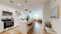 This well-designed 2-bedroom, 3-bathroom residence in Austin, TX offers a thoughtful open-concept layout that maximizes every square foot. The kitchen is a standout, featuring crisp white shaker cabinets, stainless steel appliances, a white quartz countertop, subway tile backsplash, and a central island with bar seating illuminated by striking gold pendant lights. The kitchen flows seamlessly into the bright living room, which is bathed in natural light from multiple windows dressed with sheer curtains and fitted blinds. Light wood-tone flooring runs throughout the main living areas, lending warmth and continuity. The primary bedroom is a comfortable retreat with carpet flooring, generous natural light from dual windows, and access to a walk-in closet featuring built-in shelving and hanging rods. The secondary bedroom mirrors the same calm, neutral aesthetic. The bathrooms are finished with white cabinetry, quartz countertops, large mirrors with vanity lighting, and tub/shower combinations clad in white tile. Three full bathrooms ensure ample convenience. Throughout the space, a cohesive palette of white walls, warm wood tones, and gold accents creates a modern yet inviting atmosphere well-suited for Austin living.