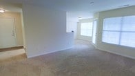 This single-floor residential space offers a clean, functional layout designed to maximize comfort and usability. The living area is finished with wall-to-wall plush carpeting in a neutral tone, creating a warm and inviting atmosphere underfoot. Soft, light-toned walls throughout lend the space a bright, airy feel that enhances the sense of openness.

A defining feature of the main living area is its abundance of natural light, drawn in through large windows fitted with blinds and a glass-paneled door that leads to an exterior access point. This combination of windows and door not only fills the room with daylight but also provides a seamless connection to the outdoors.

A half-wall partition subtly separates the living space from an adjacent area, adding architectural interest while maintaining an open-concept flow. A built-in ledge or countertop detail is visible atop this partition, offering a practical surface element. Overhead, a flush-mount ceiling light fixture provides even illumination throughout the space.

The overall ambiance is calm and neutral, offering a versatile backdrop well-suited for a variety of furnishing styles. The space presents itself in a clean, move-in-ready condition, making it an approachable and functional single-level living environment.