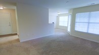This single-floor residential space offers a clean, functional layout designed to maximize comfort and usability. The living area is finished with wall-to-wall plush carpeting in a neutral tone, creating a warm and inviting atmosphere underfoot. Soft, light-toned walls throughout lend the space a bright, airy feel that enhances the sense of openness.

A defining feature of the main living area is its abundance of natural light, drawn in through large windows fitted with blinds and a glass-paneled door that leads to an exterior access point. This combination of windows and door not only fills the room with daylight but also provides a seamless connection to the outdoors.

A half-wall partition subtly separates the living space from an adjacent area, adding architectural interest while maintaining an open-concept flow. A built-in ledge or countertop detail is visible atop this partition, offering a practical surface element. Overhead, a flush-mount ceiling light fixture provides even illumination throughout the space.

The overall ambiance is calm and neutral, offering a versatile backdrop well-suited for a variety of furnishing styles. The space presents itself in a clean, move-in-ready condition, making it an approachable and functional single-level living environment.