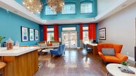 Located at 6233 Dougherty Rd in Dublin, CA, this single-floor leasing office makes a bold first impression with a striking interior design palette of teal, orange, and red. The main reception area features soaring double-height ceilings lined with oversized windows that flood the space with natural light, complemented by dramatic sculptural chandeliers overhead. Rich hardwood flooring runs throughout, anchoring the vibrant color scheme with warmth and continuity.

The lounge area is thoughtfully furnished with a mix of blue accent chairs, a neutral sectional sofa adorned with orange throw pillows, and a sleek round coffee table — all arranged to encourage comfortable, informal conversation. A trio of framed abstract artwork adorns the teal accent wall, adding a curated, gallery-like feel to the seating zone.

A dedicated workstation area includes computer desks and ergonomic seating, providing a functional space for leasing consultations. Bold red curtains frame the large glass entry doors and windows, creating a vivid contrast against the teal walls and drawing the eye toward the bright outdoor views beyond.

A bar-style counter with upholstered stools serves as a secondary engagement point, while a separate staff workspace features warm orange accent walls, built-in cabinetry, and ample desk space. The overall ambiance is energetic yet welcoming, reflecting a modern and design-forward professional environment.