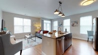 This single-floor model unit showcases an open-concept layout that seamlessly connects the kitchen and living area, creating a spacious and cohesive living environment. The kitchen serves as the centerpiece of the space, featuring a large island with granite countertops, an undermount sink, dark cabinetry, and stainless steel appliances, including a built-in dishwasher. Stylish industrial pendant lighting hangs above the island, adding a contemporary design touch.

The living area flows naturally from the kitchen, bathed in natural light streaming through multiple large windows dressed with horizontal blinds and floor-length curtains. The neutral gray walls and light hardwood-style flooring provide a clean, modern backdrop throughout the space. Recessed lighting in the ceiling complements the natural brightness of the room.

The overall ambiance is modern and inviting, with a well-considered design that balances functionality and style. The open layout maximizes the sense of space, making it feel airy and connected, ideal for both everyday living and entertaining.