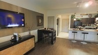 This well-appointed single-floor residence showcases a thoughtfully designed open-concept layout that seamlessly connects the main living areas. The dining space is anchored by a dark-toned bistro-height table with matching bar-style chairs, creating an intimate yet functional setting for everyday meals or entertaining. Rich, warm hardwood flooring grounds the dining and living areas, while light-colored tile extends naturally into the hallway and kitchen, providing a smooth visual transition between spaces.

The walls feature a sophisticated two-tone treatment, with a warm taupe accent wall complementing the neutral surrounding tones and adding depth to the space. Decorative artwork and carefully placed accent pieces, including a sculptural dried floral arrangement atop a textured credenza, contribute to the refined ambiance throughout.

The kitchen is visible from the dining area and is outfitted with light cabinetry, a raised breakfast bar with sleek modern stools, and stainless steel appliances, including a full-size refrigerator. A pendant light fixture adds a stylish focal point above the kitchen area. A hallway beyond the dining space leads to additional rooms, with arched doorways and clean architectural lines reinforcing the cohesive and elegant design of the interior.