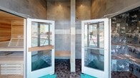 This contemporary wellness space is thoughtfully designed to deliver a full relaxation and recovery experience. The facility is anchored by a cedar-lined sauna, fully outfitted with tiered wooden benches and smooth tongue-and-groove paneling that creates a warm, aromatic environment ideal for heat therapy. Adjacent to the sauna is a dedicated steam room, its walls and built-in bench covered floor to ceiling in striking graffiti-inspired tile art rendered in tones of teal, white, and gray — a bold artistic statement that gives the space a distinctly urban aesthetic. Rinse showers are positioned between the wellness rooms, featuring large-format gray wall tiles, dark hexagonal mosaic flooring, and glass door enclosures that maintain an open, airy feel. Teal accent mats punctuate the dark tile floors throughout, adding a pop of color that ties the design together. Floor-to-ceiling glass doors and large windows flood the transition areas with natural light and offer direct views onto an outdoor pool deck, seamlessly connecting the interior wellness experience with the exterior amenity space. The overall design balances industrial-modern architecture with artful detailing, creating a space that is both functional and visually engaging.