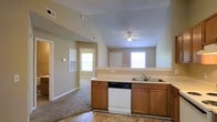This single-floor space features an open-concept layout that seamlessly connects the kitchen and living area, creating a spacious and functional environment. The kitchen is equipped with warm wood-toned cabinetry, a double-basin sink, a white dishwasher, and light countertops that extend along a peninsula, providing a natural division between the kitchen and the adjoining living space. The flooring in the kitchen area consists of tile, while the living and adjacent spaces transition to carpeting, adding a layer of comfort underfoot. Vaulted ceilings rise above the main living area, contributing to an airy, open feel, and a ceiling fan is centrally positioned to aid in air circulation. Multiple windows allow natural light to filter throughout the space, brightening the neutral beige walls. A hallway visible from the main area leads to additional rooms, suggesting a well-organized floor plan. The overall ambiance is practical and welcoming, with a straightforward design that prioritizes functionality and ease of movement throughout the connected living spaces.