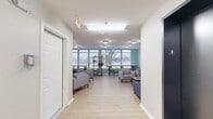 Located at 1016 McGowan Dr in Little Rock, Arkansas, this single-floor communal space is thoughtfully designed to serve as a welcoming gathering and relaxation area. The layout is divided into two distinct zones that flow seamlessly into one another, offering a variety of seating and activity options throughout.

The main lounge area features light hardwood-style flooring, crisp white walls, and flush-mount ceiling lighting that keeps the space bright and inviting. Comfortable gray and blue upholstered sofas and armchairs are arranged around low coffee tables, creating casual conversation and relaxation nooks. A wall-mounted flat-screen television anchors one seating area, while decorative touches such as a world map wall art and small potted plants add personality to the space.

The windowed lounge section is highlighted by a teal accent wall and a generous bank of windows that frame outdoor views of surrounding greenery, flooding the area with natural light. A long bar-height table with stools and open metal shelving provide functional spaces for studying or casual dining.

A wide corridor connects both areas, lined with additional seating and clear pathways to access doors. The overall ambiance is clean, modern, and functional — well-suited for community interaction, study, or quiet relaxation.