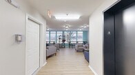 Located at 1016 McGowan Dr in Little Rock, Arkansas, this single-floor communal space is thoughtfully designed to serve as a welcoming gathering and relaxation area. The layout is divided into two distinct zones that flow seamlessly into one another, offering a variety of seating and activity options throughout.

The main lounge area features light hardwood-style flooring, crisp white walls, and flush-mount ceiling lighting that keeps the space bright and inviting. Comfortable gray and blue upholstered sofas and armchairs are arranged around low coffee tables, creating casual conversation and relaxation nooks. A wall-mounted flat-screen television anchors one seating area, while decorative touches such as a world map wall art and small potted plants add personality to the space.

The windowed lounge section is highlighted by a teal accent wall and a generous bank of windows that frame outdoor views of surrounding greenery, flooding the area with natural light. A long bar-height table with stools and open metal shelving provide functional spaces for studying or casual dining.

A wide corridor connects both areas, lined with additional seating and clear pathways to access doors. The overall ambiance is clean, modern, and functional — well-suited for community interaction, study, or quiet relaxation.