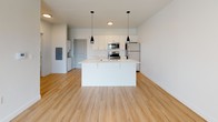 This single-floor unit features a clean, modern design highlighted by light hardwood-style flooring that flows throughout the main living areas, creating a bright and cohesive aesthetic. The open-concept kitchen anchors the space with white flat-panel cabinetry, stainless steel appliances including a range, microwave, and refrigerator, and a central island with a built-in sink and light countertop surface. Two sleek black pendant lights hang above the island, adding a contemporary touch. The kitchen opens directly into a generously sized living area, allowing for flexible furniture arrangements and an airy, connected feel. A washer and dryer unit is conveniently tucked near the entryway. The bedroom is finished with wall-to-wall carpet, offering a softer, quieter environment, and features neutral white walls that provide a calm, versatile backdrop. The bathroom is appointed with light hardwood-style flooring, a white vanity with an integrated sink, a large mirror, and a full-sized soaking tub with a shower. Throughout the unit, the consistent use of white walls and light flooring contributes to a bright, clean atmosphere well-suited to modern living.