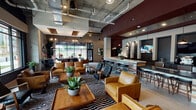 Located at 1415 Iris Drive in Charlotte, NC, this single-floor space presents a thoughtfully designed communal lounge that blends industrial and contemporary aesthetics. The open-plan layout is anchored by exposed concrete columns, visible ductwork overhead, and polished concrete floors, creating an urban, loft-inspired atmosphere throughout.

The main gathering area features generous lounge seating, including tan leather sofas and armchairs arranged around bold black-and-white striped area rugs, complemented by live tropical plants and curated artwork on deep charcoal accent walls. A prominent bar and counter area with barstool seating, pendant lighting, and what appears to be a beverage station serves as a social focal point, enhanced by mounted televisions and decorative tile detailing.

Floor-to-ceiling black-framed windows span multiple walls, flooding the interior with abundant natural light and offering clear views of the surrounding streetscape and landscaped exterior. A separate work-friendly zone provides communal table seating with task-appropriate lighting, ideal for collaborative use.

Distinctive pendant and cage-style light fixtures, concrete structural elements, and carefully placed greenery tie the industrial framework to a warm, welcoming ambiance throughout the space.