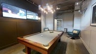 This single-floor entertainment lounge is designed with a modern, industrial aesthetic, creating an inviting atmosphere for recreation and socializing. The centerpiece of the space is a full-size pool table with a light gray felt surface and rich wood finish, positioned beneath a striking geometric cluster chandelier fitted with adjustable track lighting that bathes the room in warm, focused illumination. The exposed ceiling with visible ductwork and mechanical elements reinforces the industrial character of the space, complemented by cool gray walls that give the room a sleek, contemporary feel. A large flat-screen television is mounted prominently on the wall, making the space well-suited for watching sports or entertainment during gatherings. Seating is provided by a pair of modern swivel lounge chairs in a muted blue-gray tone, offering comfortable spots to relax between games. Textured, patterned carpeting covers the floor, adding warmth and visual interest to the otherwise industrial design. A glass-paneled door provides access to an adjacent area, enhancing the sense of openness. The overall ambiance is energetic yet comfortable, combining functional amenities with thoughtful design to create a well-appointed communal recreation space.