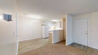 This single-floor space showcases an open-concept design that seamlessly connects the living area and kitchen. The living room features plush carpet flooring and neutral-toned walls, creating a warm and inviting atmosphere. A decorative chandelier serves as a focal point overhead, adding a touch of elegance to the space. The textured ceiling extends throughout, maintaining a consistent aesthetic.

The kitchen is positioned at the rear of the open layout and is defined by a peninsula-style counter with light wood-toned cabinetry below, offering both workspace and a natural divider between the two areas. White upper cabinets provide ample storage, while a full-size white refrigerator and built-in sink round out the functional kitchen space. Overhead fluorescent lighting ensures the kitchen area is well-lit.

Light hardwood-style flooring in the kitchen transitions smoothly into the carpeted living area, subtly distinguishing the two zones while maintaining an open flow. The overall color palette of whites and warm beiges contributes to a clean, bright, and cohesive environment throughout the space.