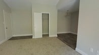This single-floor space offers a clean, functional layout with a neutral color palette that creates a calm and welcoming atmosphere throughout. The interior is finished with soft beige walls and white trim baseboards, lending a fresh and well-maintained appearance.

The bedroom area is finished with plush, light-colored carpeting that adds warmth and comfort underfoot. A built-in sliding-door closet provides convenient storage, featuring a clothes rod and open shelving within. The room's simple, understated design allows for flexible furniture arrangements.

Adjacent to the bedroom, the flooring transitions seamlessly to rich, dark hardwood planks, defining a separate living or common area. This zone is accented by a pendant light fixture with a frosted glass shade that hangs from the ceiling, providing both ambient illumination and a subtle decorative touch.

The consistent neutral wall tones and white trim throughout both areas create a cohesive aesthetic that is easy to personalize. The straightforward, practical design of this space makes it well-suited for a variety of uses, offering a solid foundation for comfortable, everyday living.