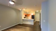 This single-floor space offers a well-designed open-concept layout that seamlessly connects the living area to the kitchen, creating a cohesive and functional living environment. The kitchen is a standout feature, boasting warm orange-toned wood cabinetry that adds a rich, inviting character to the space. A peninsula with a light-colored countertop provides both additional prep space and a natural divider between the kitchen and living area, making it ideal for casual dining or entertaining. A classic tile backsplash adds texture and visual interest, while black appliances, including a refrigerator, microwave, and range, lend a sleek, modern contrast to the warm cabinetry. Track lighting runs along the ceiling of the kitchen area, ensuring the workspace is well-lit and functional. The living area benefits from the same rich, dark hardwood-style flooring that flows throughout the unit, grounding the space with warmth and continuity. Neutral gray-toned walls provide a versatile backdrop that complements a variety of décor styles. A hallway leads toward what appears to be additional rooms, including a closeted space, suggesting a practical and efficient floor plan. The overall ambiance is modern, warm, and move-in ready.